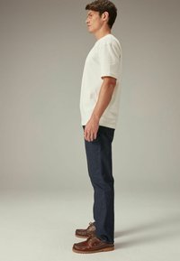Witte t-shirt met korte mouwen, donkerblauwe spijkerbroek en bruine leren schoenen met veters. De outfit is vanaf de zijkant gezien tegen een neutrale achtergrond.
