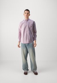 Camisa de botones en rosa claro con rayas blancas, combinada con jeans azul holgados y zapatillas marrones. Cuenta con un bolsillo en el pecho y mangas largas.
