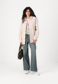 Giacca oversize beige chiaro, t-shirt con grafica, jeans a zampa d'elefante, sneakers bianche e borsa a tracolla verde. Texture morbida e vestibilità rilassata.