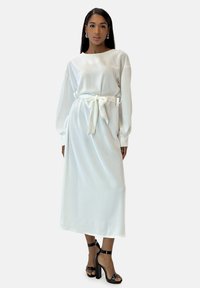 Robe longue en satin blanc à manches longues avec ceinture à la taille, dotée d'un col rond et d'une jupe fluide. Le modèle porte des sandales à talons noires.