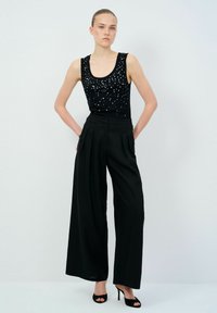 Zwarte tanktop met zilveren versieringen, gecombineerd met hoge taille wijde zwarte broeken en zwarte stiletto's. Effen achtergrond.