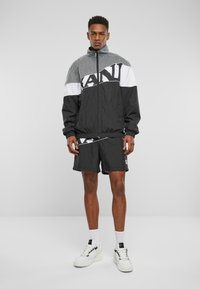 Black and white windbreaker jacket with zip closure and mesh upper, paired with matching shorts. Model wears white sneakers and ankle socks.