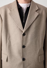 Blazer beige con un design strutturato, due tasche frontali, tre bottoni neri e un colletto a risvolto sopra un top nero con zip.