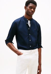 Camisa azul marino abotonada con cuello mandarín, con botones blancos y mangas remangadas. Combinada con jeans blancos, resaltando el contraste y el ajuste.