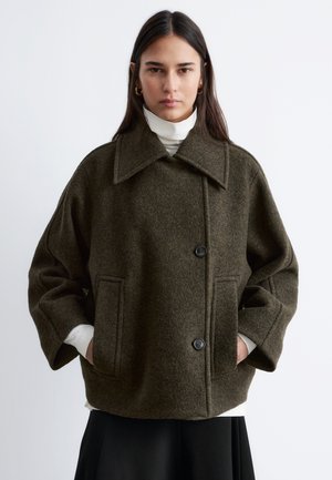 Woman with long dark hair wearing gold hoop earrings, an olive green wool coat with large collar, black buttons, and side pockets over a white turtleneck.