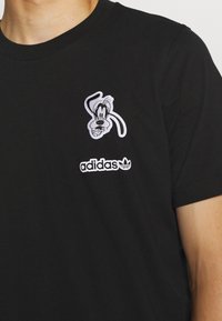 Svart bomulls T-shirt med en broderad Goofy-karaktär i vitt på vänster bröst och en "adidas"-logotyp nedanför i vitt med en ikon.