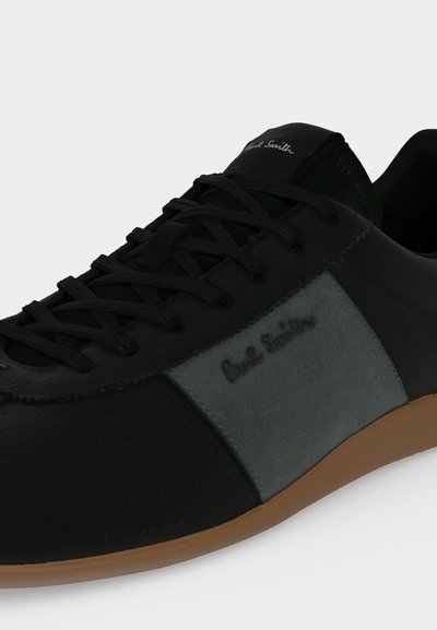 Baskets noires et grises avec lacets noirs et semelle marron, présentant la marque "Paul Smith" sur le côté et la languette.