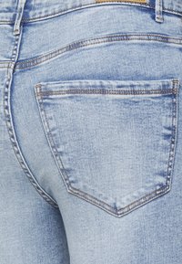 Calças de ganga azul claras com uma textura desgastada, apresentando um único bolso traseiro com costura em contraste e um detalhe de costura em ângulo no yoke.