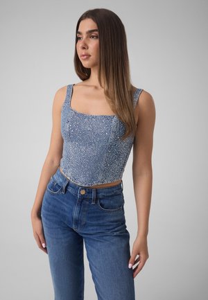 Joven con cabello largo y castaño que lleva una blusa sin mangas azul con estampado y vaqueros azules, de pie contra un fondo gris liso.