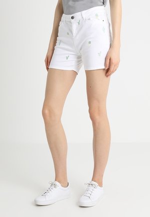 Kvinde iført hvide shorts med små grønne kaktusprints og hvide sneakers, stående mod en ensfarvet lys baggrund.