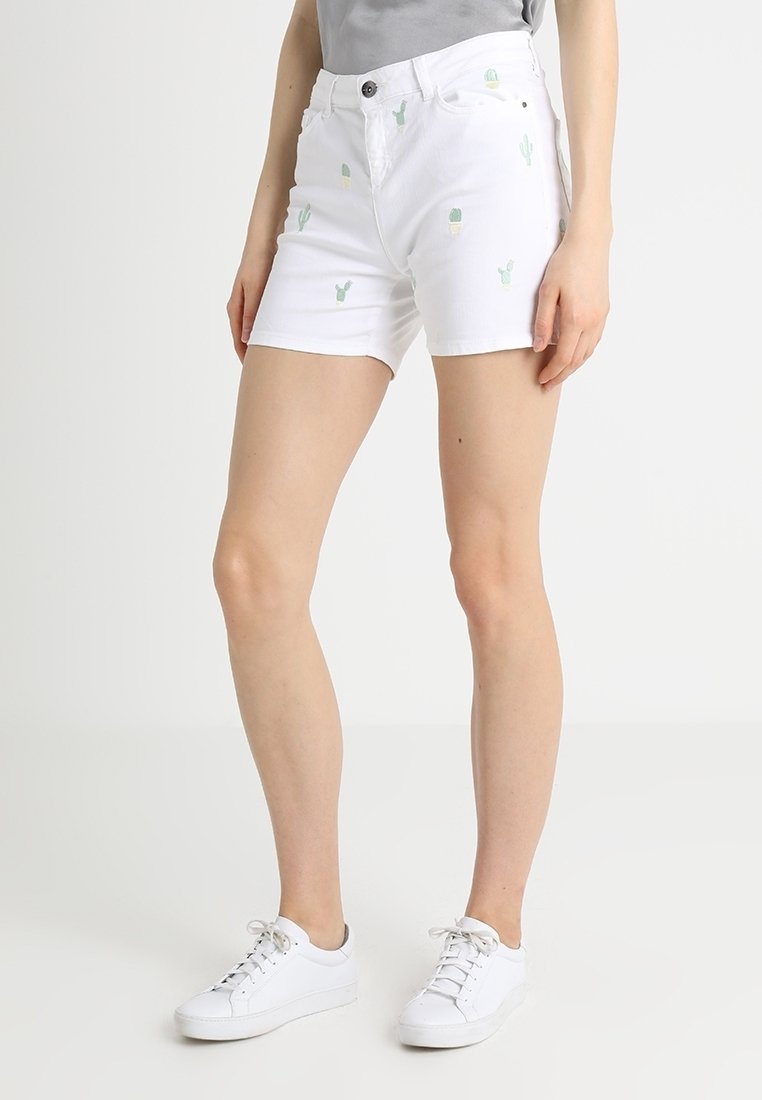 Femme portant un short blanc avec de petits imprimés cactus verts et des baskets blanches, debout devant un fond clair uni.