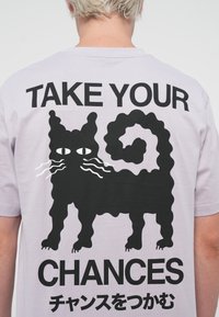 Lilla t-shirt med et sort, bølgende kattegrafik og teksten "TAG DINE CHANCER" i fede, sorte versaler.