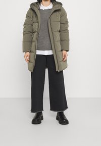 Olivengrüner gepufferter Mantel mit Kapuze, Reißverschluss und Seitentaschen, getragen über einem grauen Pullover und einem weißen Hemd mit schwarzen weit geschnittenen Hosen und Stiefeln.