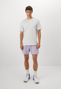 Lys grå kortærmet t-shirt kombineret med lavendelshorts. Modellen har hvide sneakers med lilla detaljer og hvide sportsstrømper på.