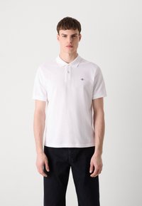 Hvit polo-shirt laget av bomull, har en klassisk krage, to knapper og en liten brodert logo på brystet. Kombinert med mørke bukser.