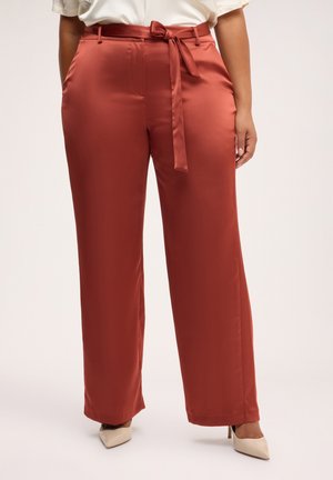Donna che indossa pantaloni in raso color ruggine a gamba larga con cintura annodata in vita e décolleté beige con punta appuntita.