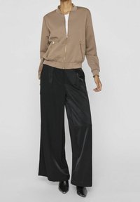 Femme portant une veste beige à fermeture éclair dorée, un pantalon large noir et des bottes noires à bout pointu, debout devant un fond uni.