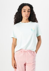 T-shirt in cotone verde menta chiaro con scollo rotondo e maniche corte. Presenta un logo colorato sul petto. Abbinato a pantaloni a righe rosa.