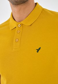 Polo-shirt jaune moutarde avec une finition texturée, doté d'une patte de boutonnage à trois boutons et d'un petit logo d'oiseau vert sur la poitrine gauche.