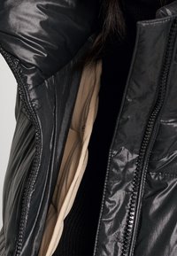 Svart vadderad jacka med en glänsande yta och beige foder. Har hel dragkedja, ribbad textur och kontrasterande inre färg.