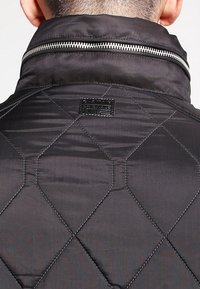 Svart vadderad jacka med hög krage, med en silverfärgad dragkedja i fronten och en vävd etikett nära kragen. Slät, syntetisk tygtextur.