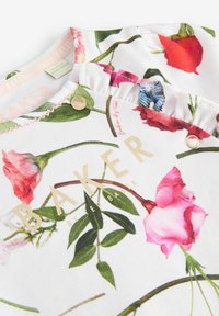 Tejido blanco con estampado floral que presenta rosas rosas y hojas verdes. Logo "BAKER" en dorado, acompañado de herrajes decorativos dorados y volantes.