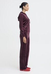 Bordowy welurowy dres z kapturem, z przednią kieszenią i długimi rękawami. Do kompletu czerwone buty wsuwane. Gładka tekstura na całej powierzchni.