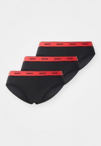 Trois slips noirs avec des ceintures élastiques rouges portant le logo "HUGO" en texte noir, disposés en pile diagonale sur fond blanc.