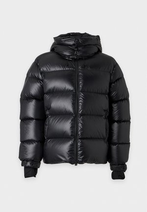Homme portant une veste matelassée noire sur une chemise blanche, un pantalon marron, une ceinture noire et des baskets noires, debout devant un fond uni.