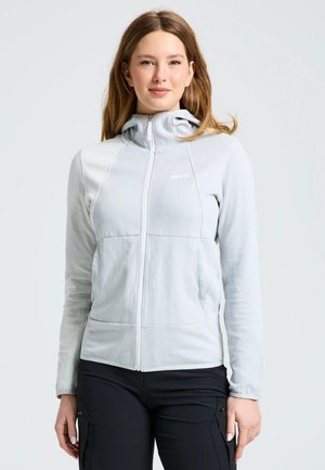 Una mujer luce una chaqueta gris claro con capucha y cremallera, junto con unos pantalones negros, sobre un fondo liso y claro.