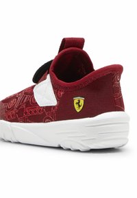 Chaussure de sport rouge avec une tige texturée, des accents blancs et une sangle noire. Elle présente un logo Ferrari jaune au niveau du talon.