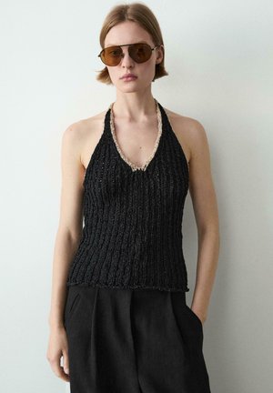 Žena s hnědými slunečními brýlemi, černým pleteným halter topem s béžovým lemováním a vysokým pasem černých kalhot, stojící u bílé stěny.