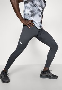 Graue Leggings mit einem strukturierten Muster, versehen mit einem weißen Nike-Logo. Kombiniert mit einem grauen, gemusterten Sportshirt und schwarzen Sneakers.