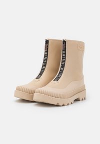 Botas de tobillo de goma beige con suelas gruesas y estriadas, cremalleras frontales con tiradores negros que llevan el texto blanco "Pepe Jeans".
