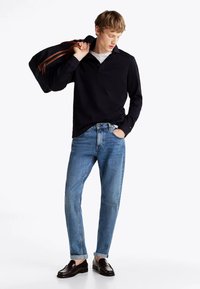 Polo a maniche lunghe blu navy, jeans in denim blu e mocassini marroni. Il modello porta una borsa nera con dettagli della chiusura marrone. Fit informale.
