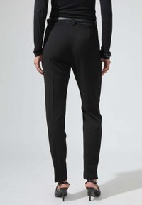 Pantalons noirs ajustés avec une coupe slim, dotés d'une texture lisse et d'une ceinture. Portés avec des talons noirs, présentant un design épuré et contemporain.