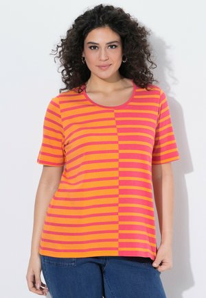 Frau mit lockigem dunklem Haar, die ein orange-pink gestreiftes T-Shirt mit kurzen Ärmeln und blaue Jeans trägt, vor weißem Hintergrund stehend.