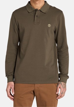 Mężczyzna ubrany w oliwkowozieloną koszulkę polo z długim rękawem z małym logo na piersi oraz brązowe spodnie, stojący z rękami swobodnie opuszczonymi wzdłuż ciała.