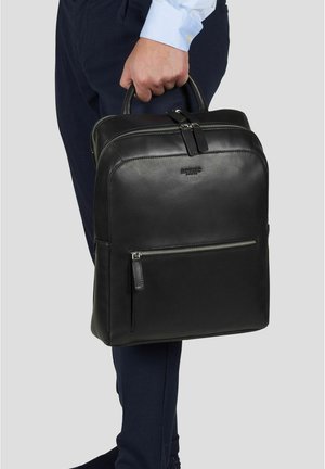 Mann i blå bukser og lyseblå skjorte som holder en svart lærmappe med frontlomme med glidelås og doble håndtak.