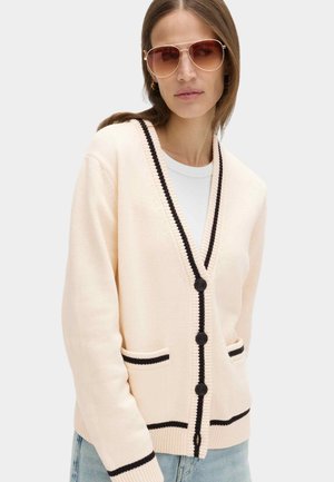 Kvinde iført cremefarvet cardigan med sort kant og knapper over hvid top, blå jeans og store pilot-solbriller.
