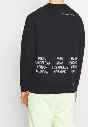 Personne portant un sweat-shirt noir avec les noms des villes en blanc et "Beaverton, Oregon" en vert imprimé dans le dos.