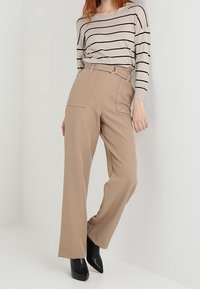 Femme portant un pantalon beige taille haute à jambes larges avec de grandes poches, un haut à rayures beige à manches longues, et des bottes pointues noires.