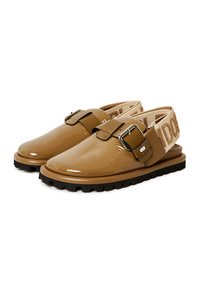 Scarpe in pelle sintetica marrone con una larga fascia e fibbia, caratterizzate da un elastico beige testurizzato e una suola spessa nera.