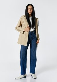 Beige waterdichte jas met een capuchon, drukknoopjes en verstelbare trekkoorden, gecombineerd met hoog gewaisted blauwe jeans en witte enkelboots.