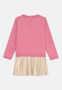 Maglia rosa a maniche lunghe con colletto arricciato, dotata di chiusura con bottone sul retro, abbinata a una gonna plissettata dorata.