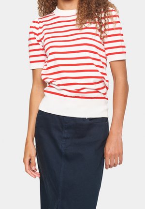 Vrouw die een wit T-shirt met korte mouwen draagt met rode horizontale strepen en een donkerblauwe rok tot op de knie tegen een witte achtergrond.