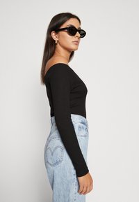 Haut noir à manches longues avec un design dénudé des épaules, associé à un jean en denim bleu clair taille haute. Les accessoires incluent des lunettes de soleil noires.