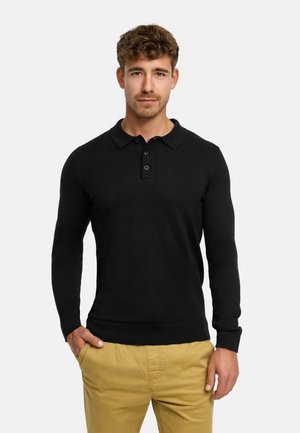 Jonge man met kort krullend haar, gekleed in een zwarte poloshirt met lange mouwen en mosterdgele broek, staand met één hand in de zak.