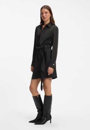 Kvinde stående iført en sort bælte-shirt kjole med lange ærmer og sorte knælange støvler med spidse tæer mod en ensfarvet hvid baggrund.