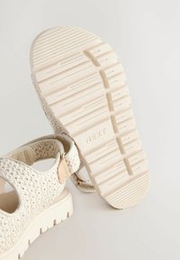 Cremfarbene, gewebte Sandalen mit einer strukturierten Oberseite, verstellbarem Riemen und einer dicken, geriffelten Gummisohle, die an der Unterseite mit "NEXT" geprägt ist.